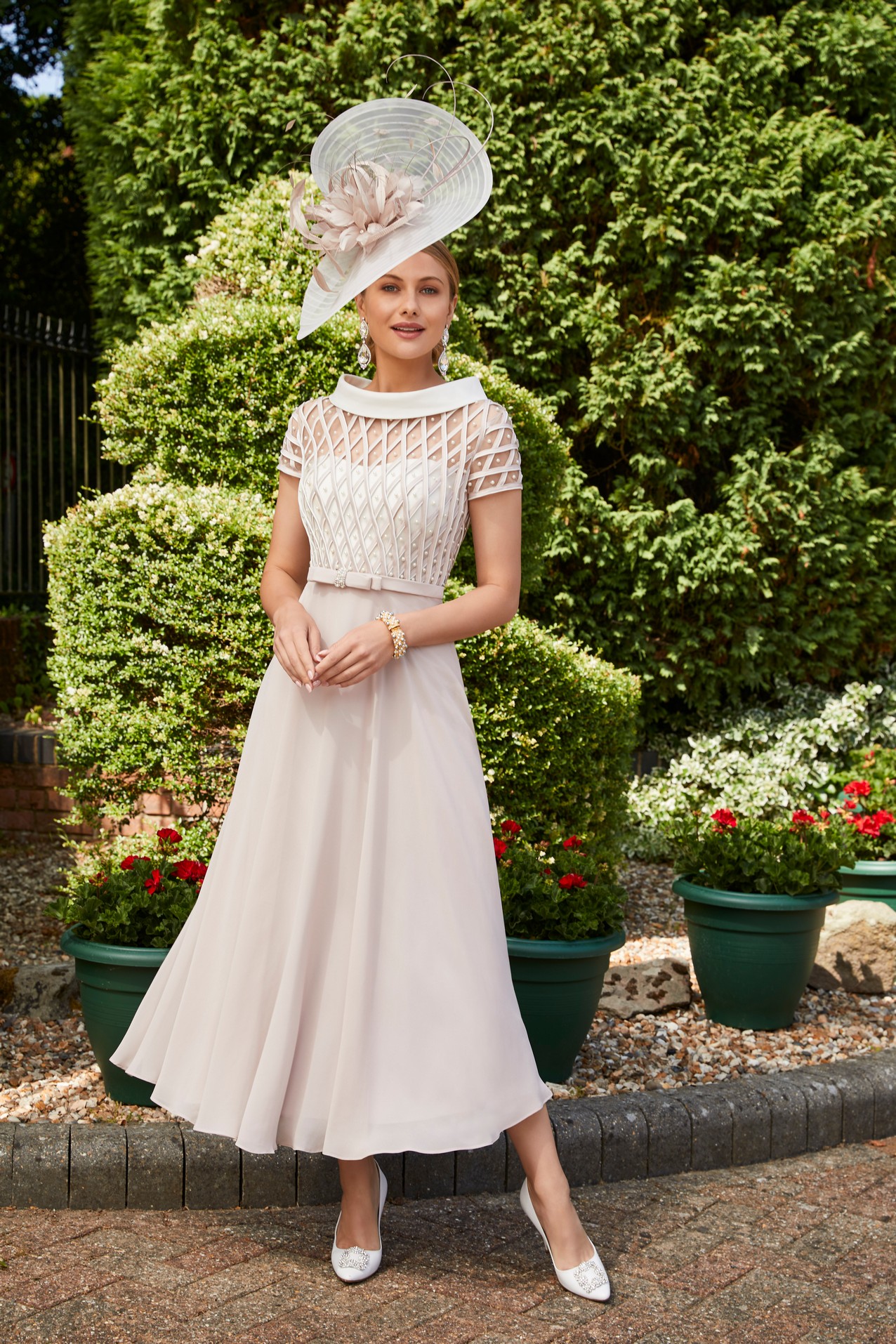 Woman standing in garden wearing nude occasion dress with pearl lattice detail and matching court heeled shoes and fascinator occasion headwear